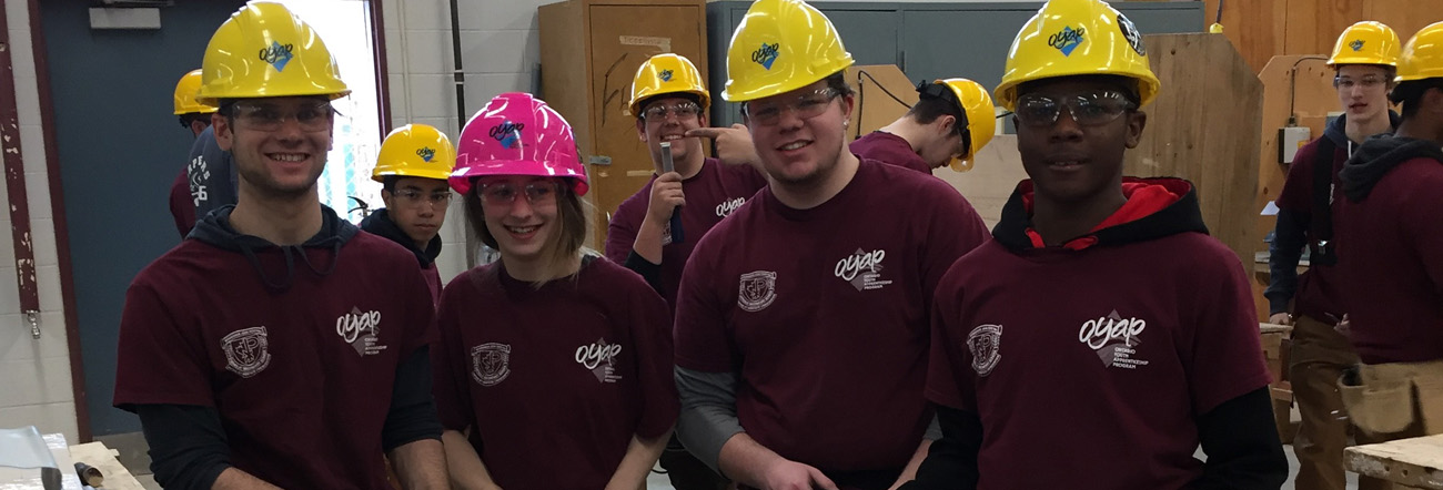 Male students wearing yellow hard hats and one female student wearing a pink hard hat in a construction classroom