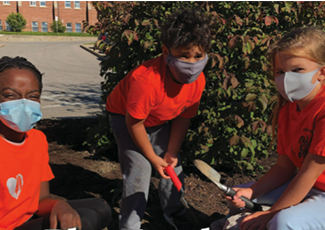 Two female and a male student wearing orange t-shirts and wearing masks outside planting in a garden